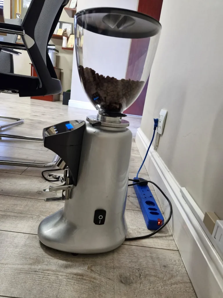 A silver Anfim on-demand espresso grinder with beans in the hopper, plugged in on a wooden floor beside office chairs in the early days of the business.