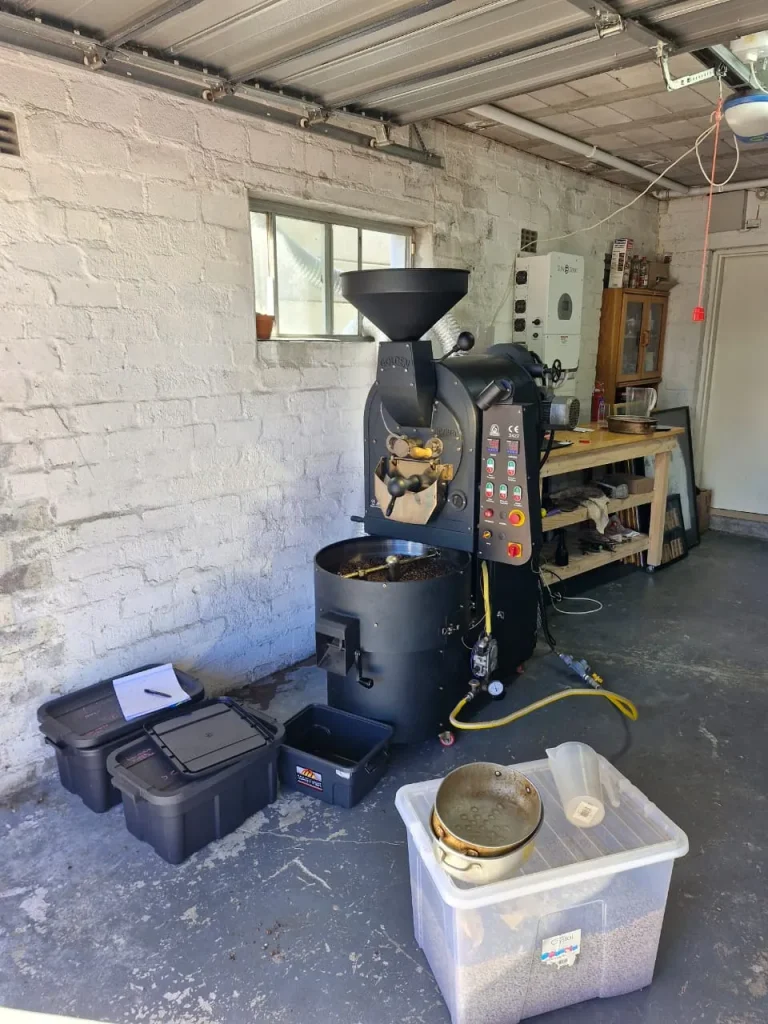 Wide view of the small roastery garage showing the black drum roaster, storage tubs, wooden workbench and brick wall