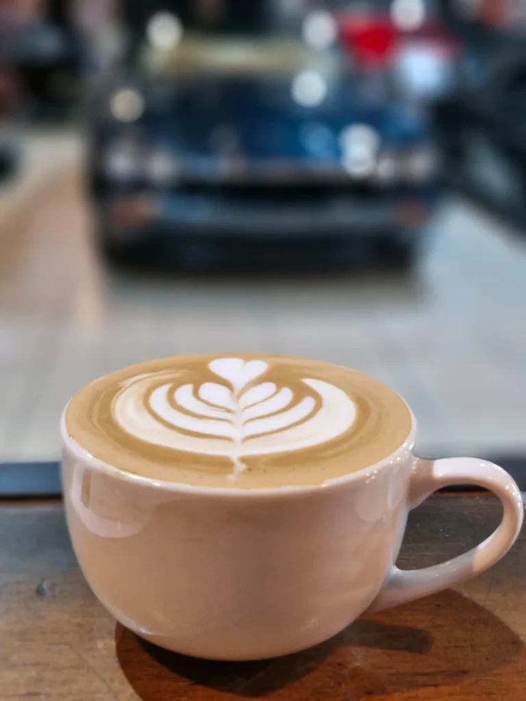 A cappuccino with fern latte art in a pale cup on a wooden ledge, a blue classic car blurred in the background at Crankshaft Cafe.