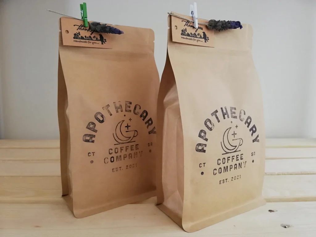 Close-up of two kraft Apothecary Coffee Company bags on a wooden surface, each finished with a 'Handmade for you' tag, a wooden peg and a sprig of dried lavender.