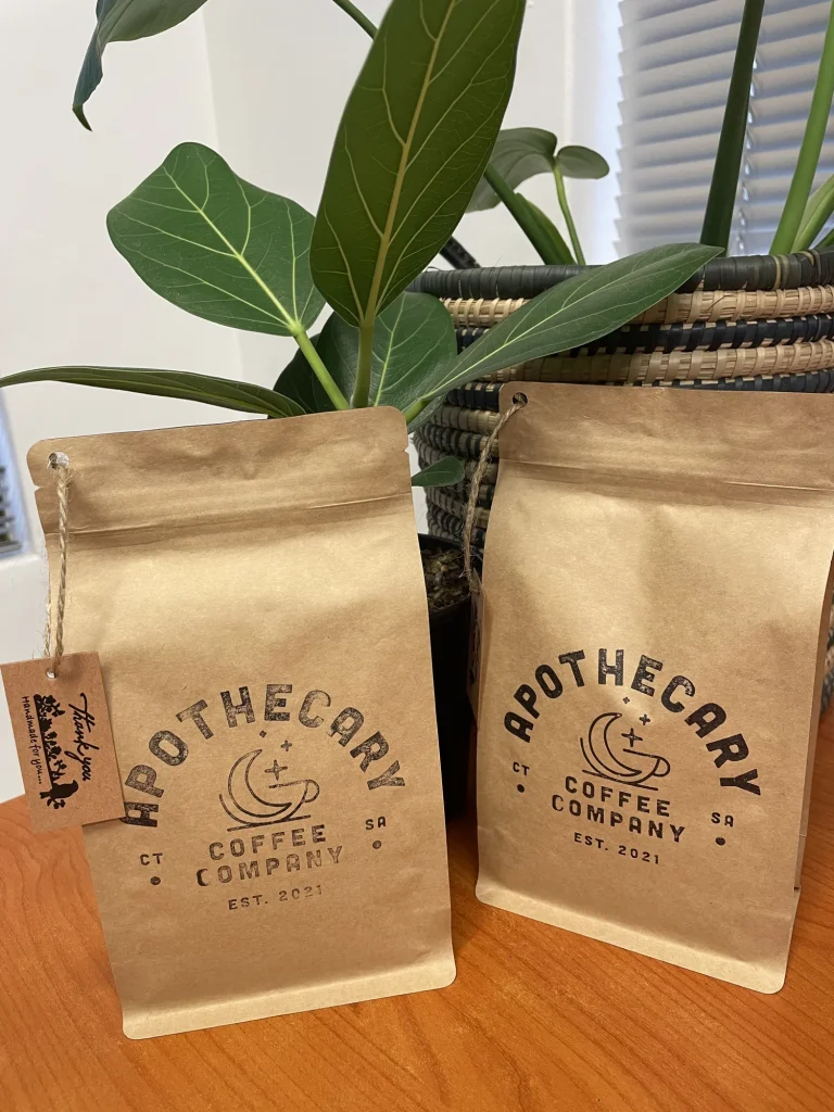 Two kraft Apothecary Coffee Company bags on a wood desk with green indoor plants and a woven basket in the background.