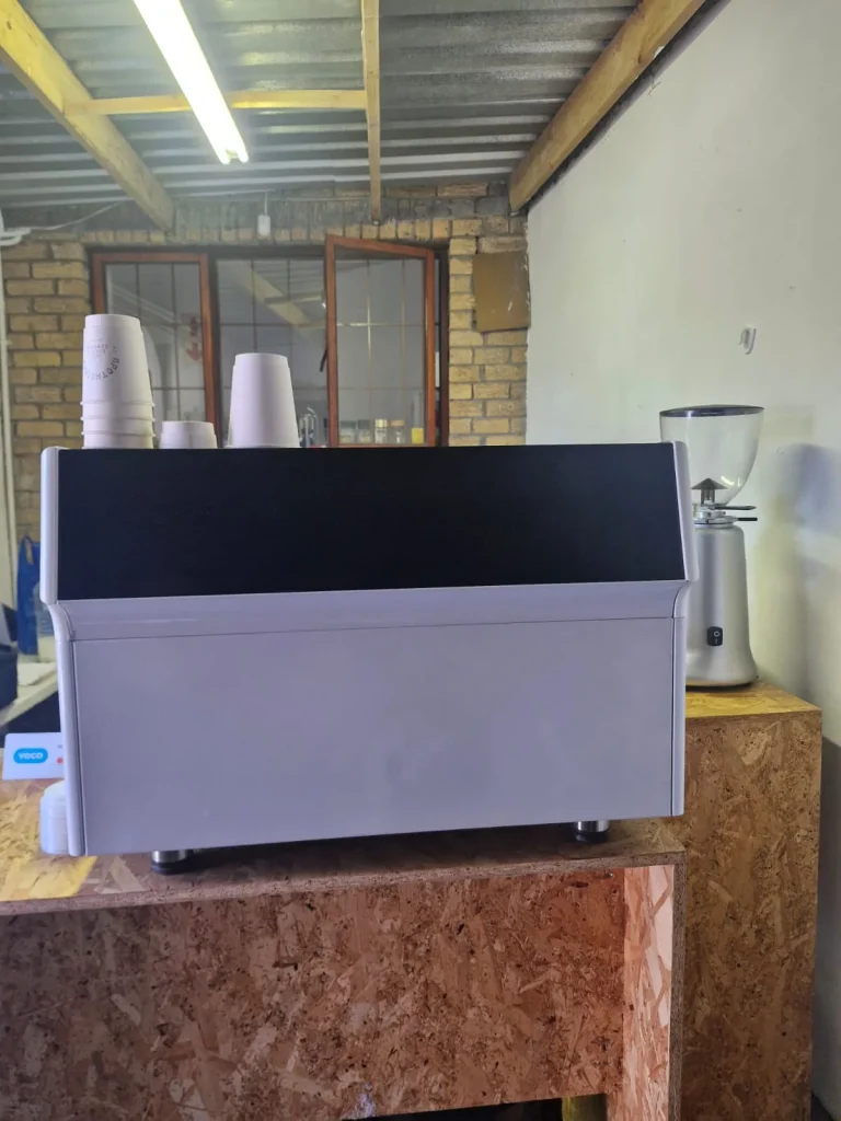 A white two-group espresso machine and grinder on an OSB timber counter inside the Apothecary Coffee roastery hatch in Blouberg, with a brick wall and Yoco card reader behind.