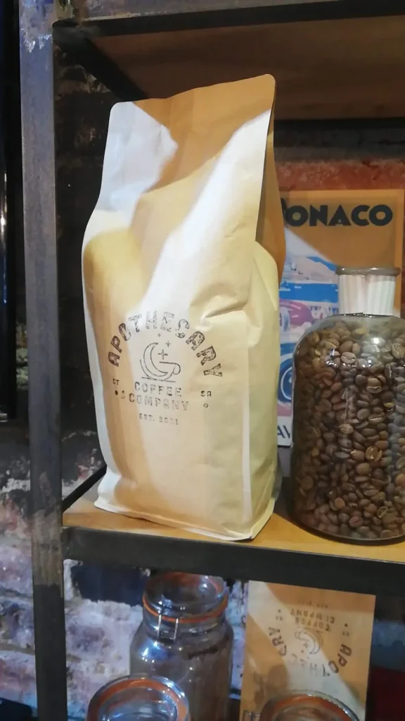 Apothecary Coffee bag standing on an industrial shelf next to a large glass jar of green coffee beans inside the Laude cafe