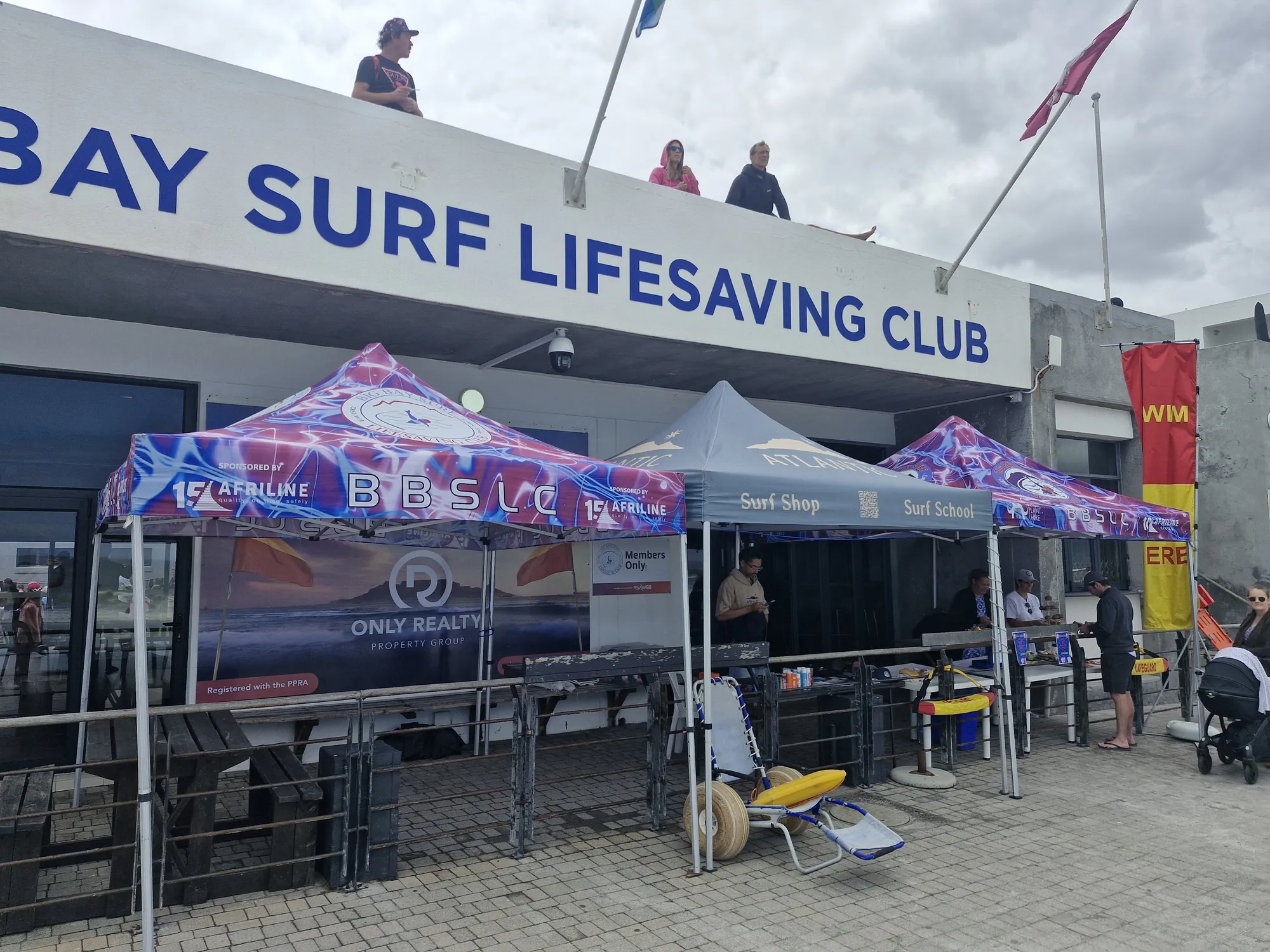 The front of the Big Bay Surf Lifesaving Club on a busy event day, with branded gazebos, flags and people gathered outside.