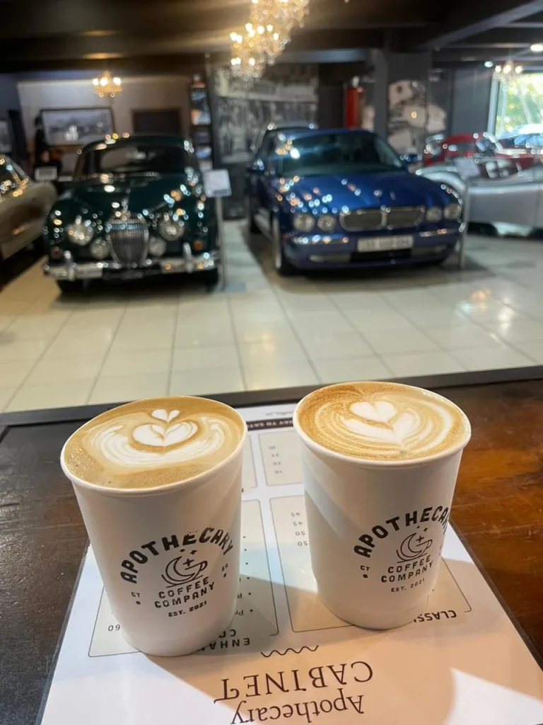 A close-up of a cappuccino with fern latte art in a pale cup, a blue classic car softly blurred in the background at Crankshaft Cafe.