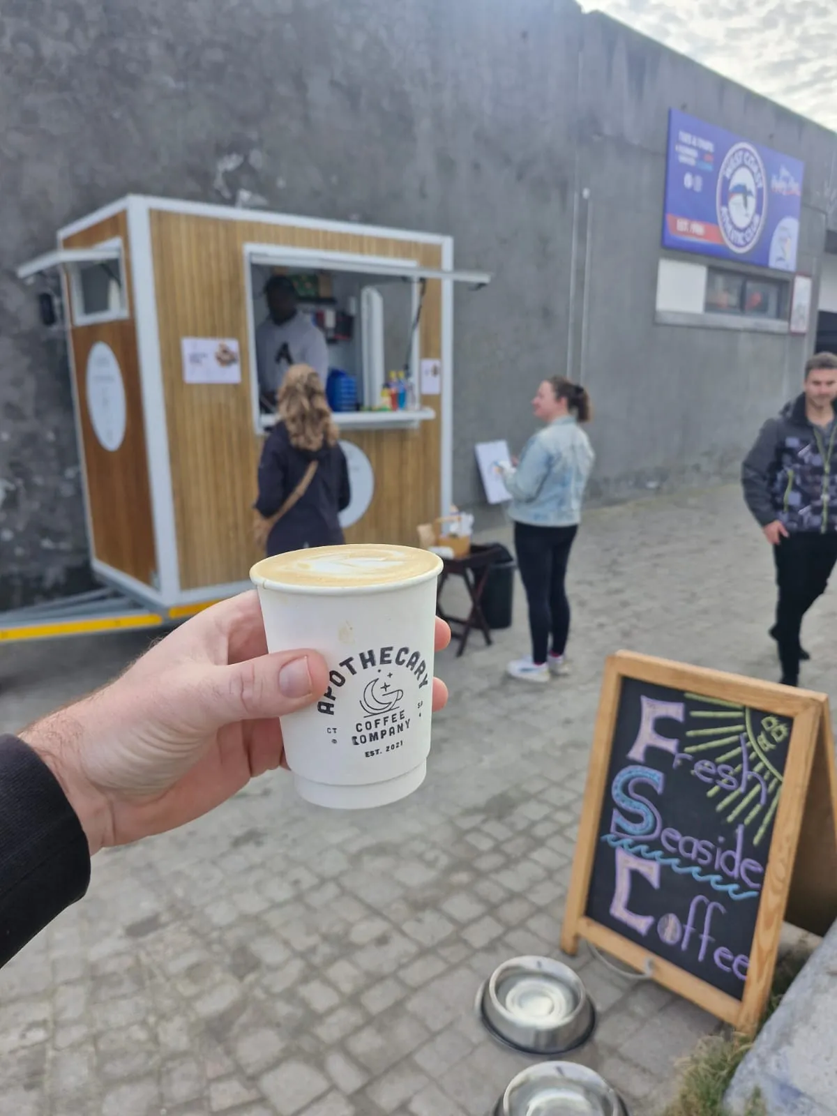 Hand holding up an Apothecary Coffee cup with heart latte art in front of the trailer at Big Bay Life Saving Club