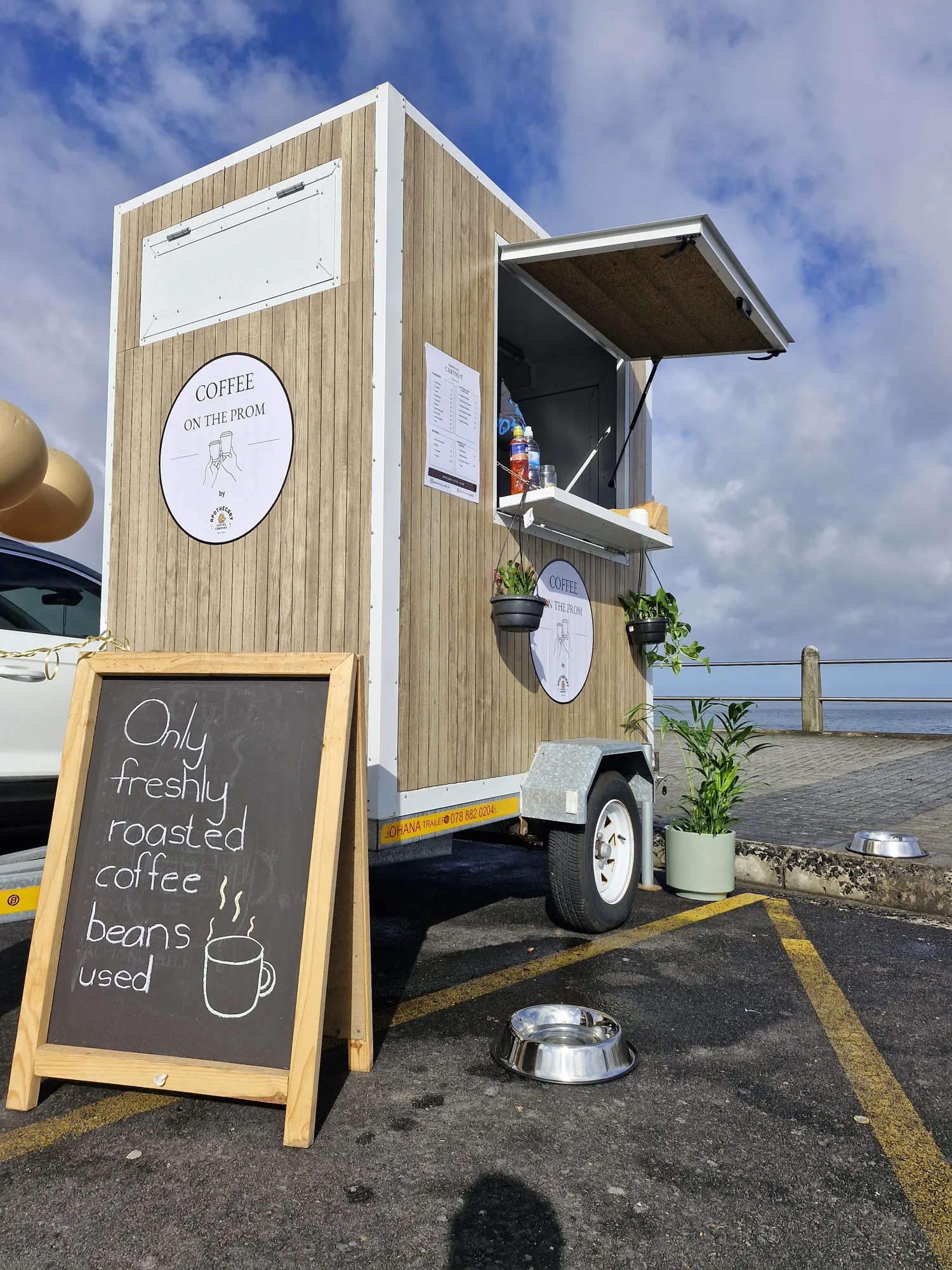A low-angle view of the wood-clad Coffee on the Prom trailer against a big blue sky at the Sea Point promenade, hatch open with a menu and dog bowls.