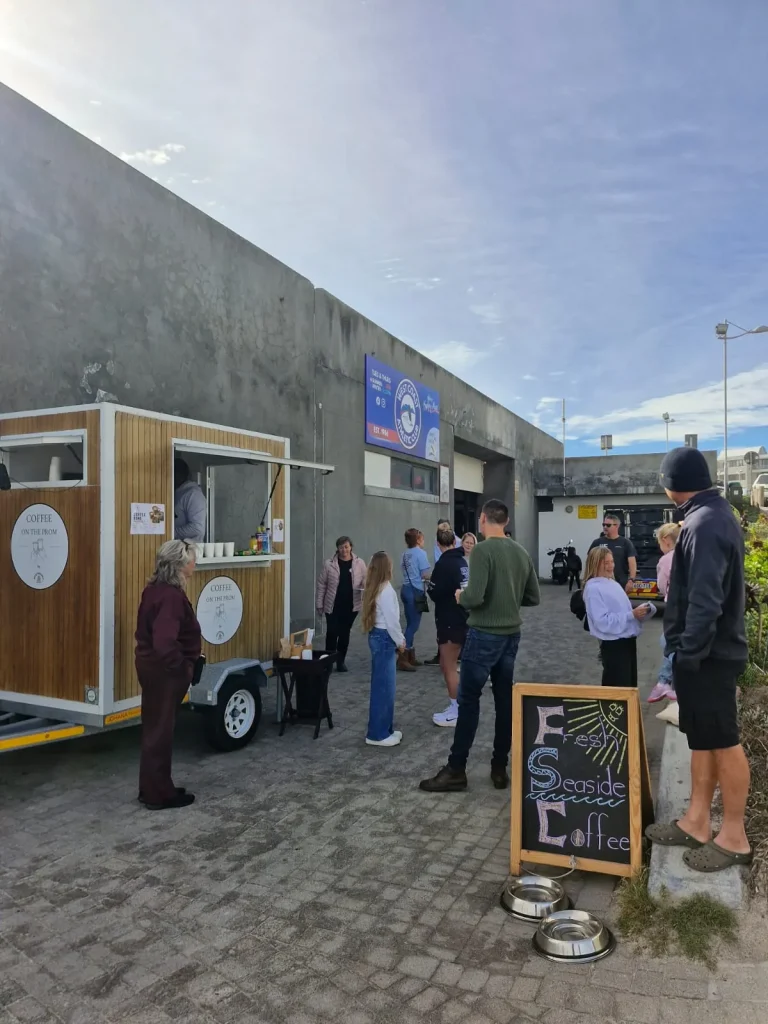 The Coffee on the Prom trailer parked outside the West Coast Athletic Club with a small queue of customers and a 'Fresh Seaside Coffee' chalkboard, on an overcast morning.