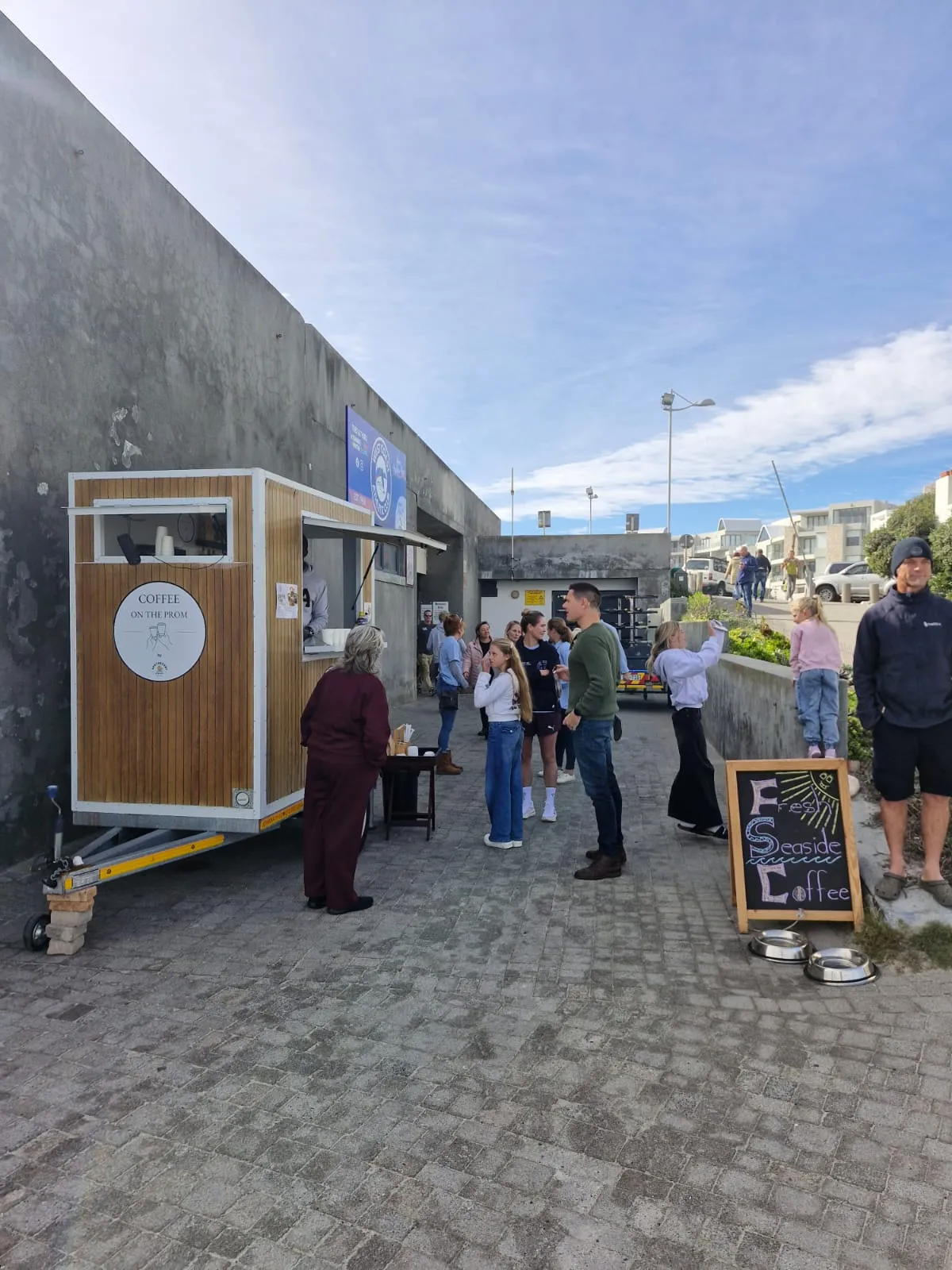 Wide shot of customers queuing at the Apothecary Coffee trailer next to the Big Bay Life Saving Club, coastal apartments in the background