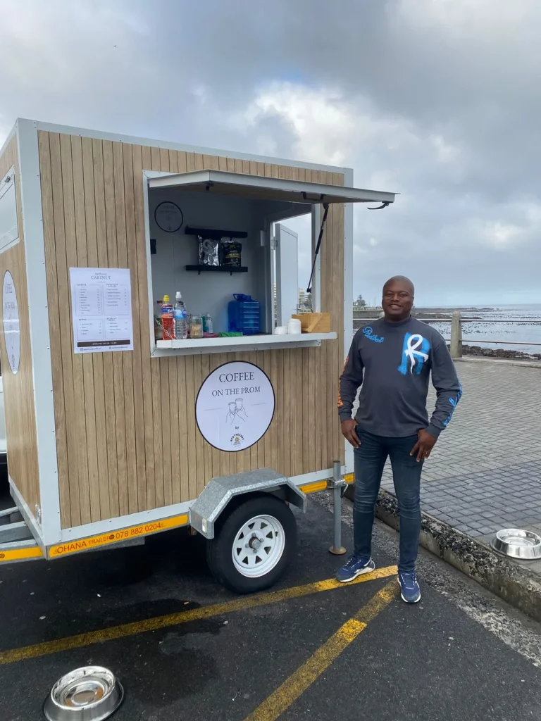 A man in a grey sweatshirt standing beside the Coffee on the Prom trailer on an overcast day at the Sea Point promenade.