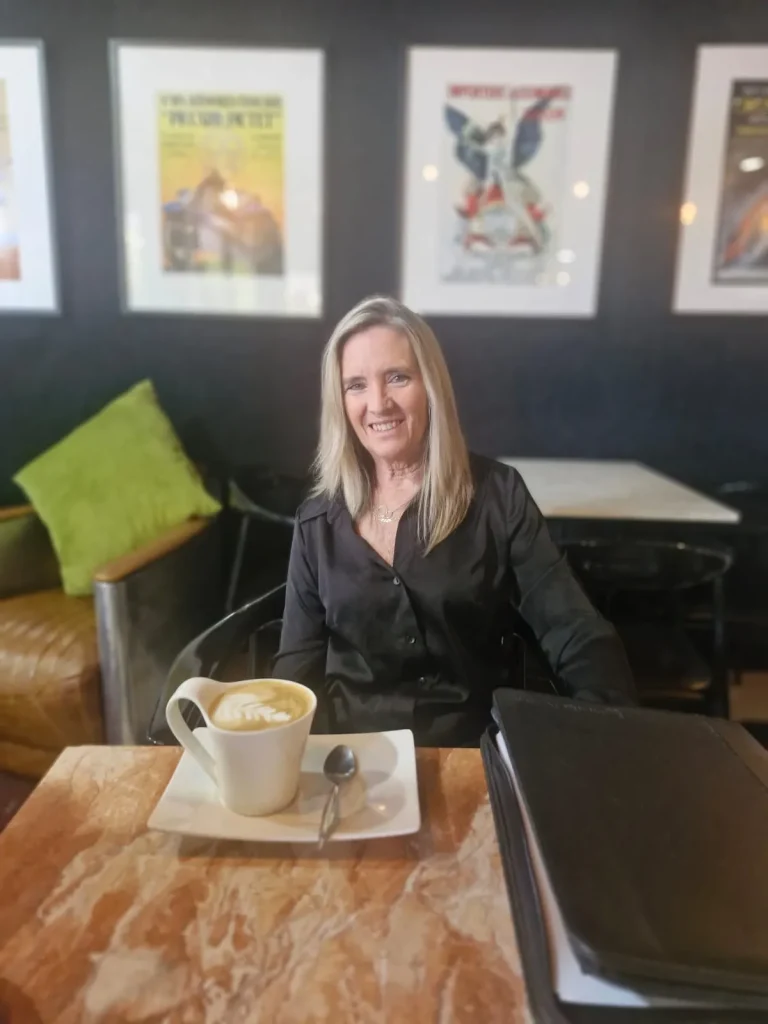 A smiling woman in black sits at a marble-topped table with a cappuccino, framed vintage motoring posters on the wall behind at Crankshaft Cafe.