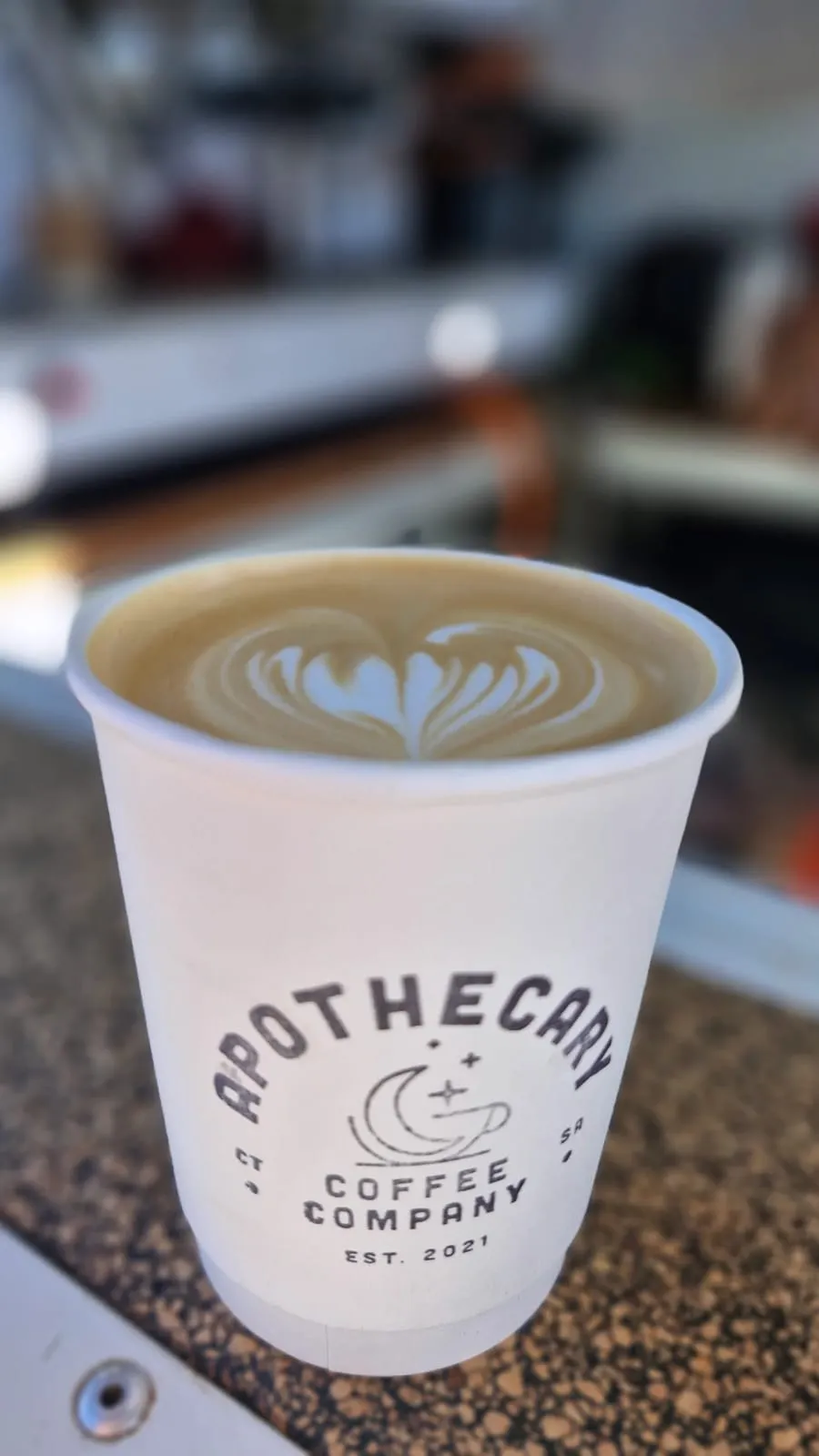 Close-up of an Apothecary Coffee takeaway cup with heart-shaped latte art, held up in front of the trailer