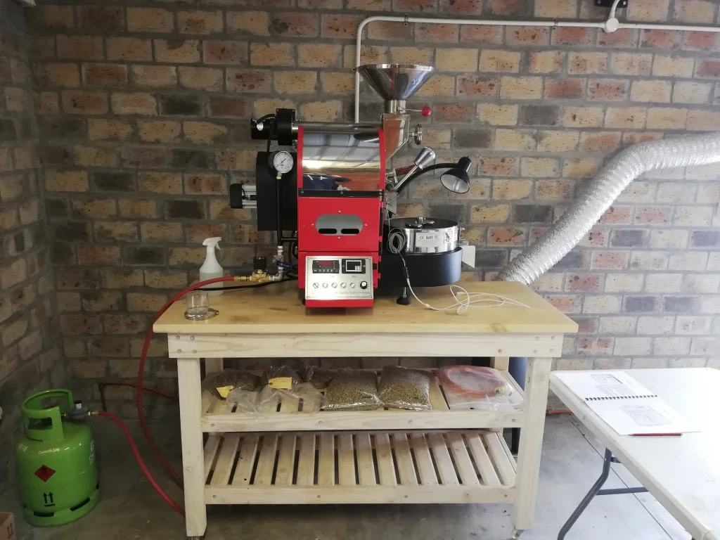 A red Mill City 1kg drum coffee roaster set up on a wooden table against a brick wall, with a gas bottle, extraction ducting and bags of green beans stored underneath.