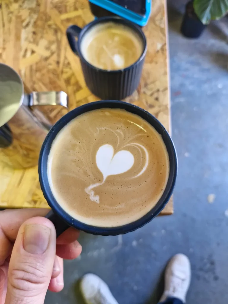 Overhead view of a flat white with heart latte art in a dark cup, held over an OSB table beside a second coffee, at the Apothecary roastery hatch.