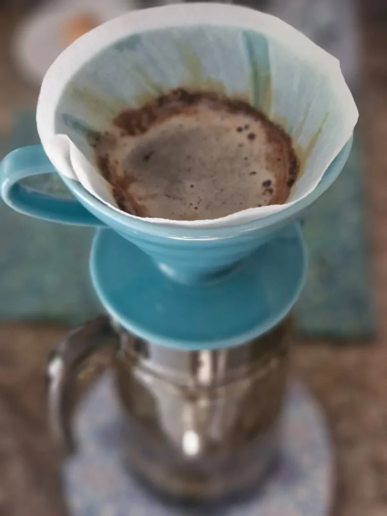Overhead shot of a teal ceramic V60 dripper with a used coffee filter and grounds, sitting on a stainless steel carafe