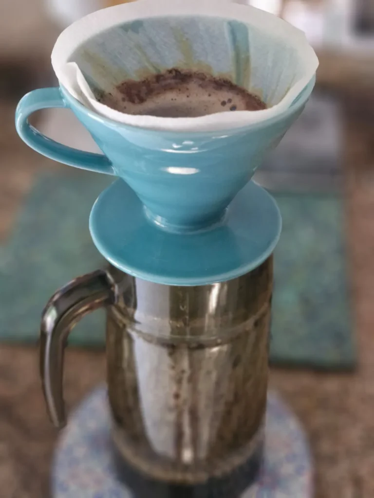 Close-up of a teal V60 dripper with coffee grounds, brewing pour-over coffee into a stainless steel carafe