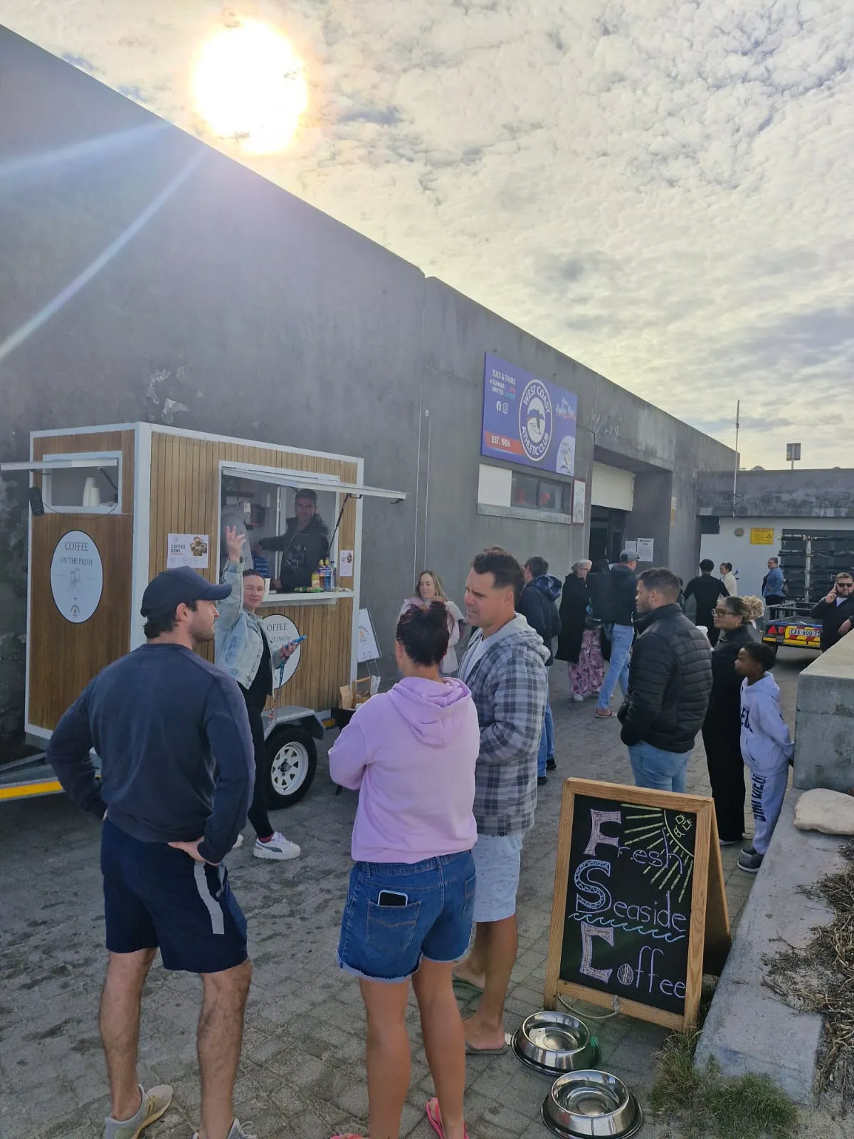 A barista serves and waves from the window of the Coffee on the Prom trailer while a crowd gathers around at a sunny sea
