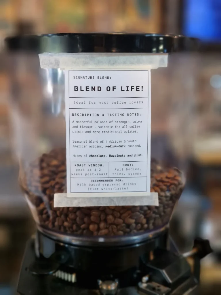 Grinder hopper filled with coffee beans, labelled Signature Blend Blend of Life with tasting notes of chocolate, hazelnut and plum