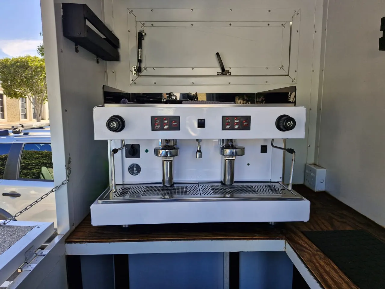 A white two-group commercial espresso machine mounted inside the Apothecary coffee trailer, viewed through the open hatch.