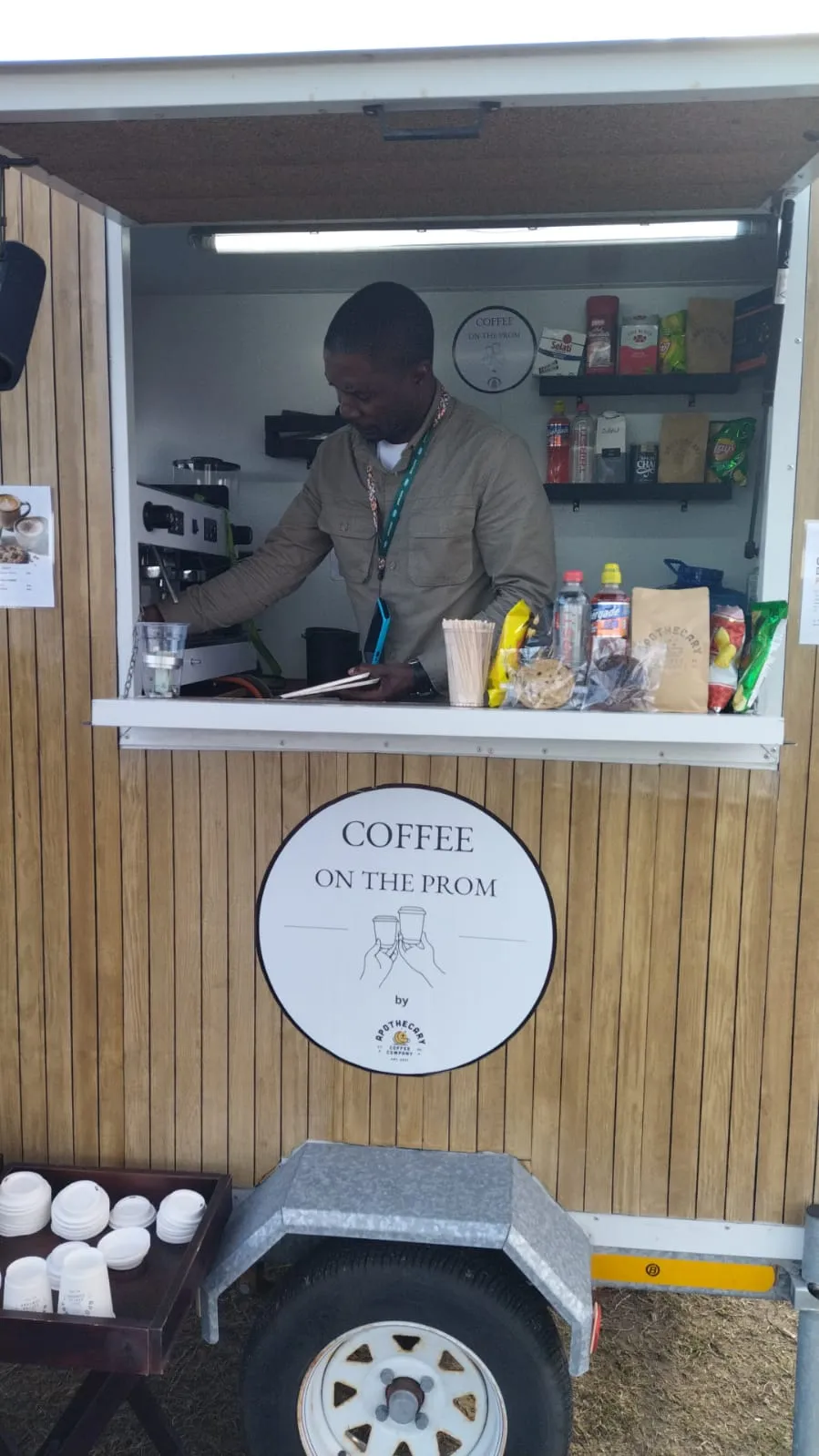 Barista Wisdom leaning out of the Apothecary Coffee trailer window, smiling, with pastries and a price list on the counter
