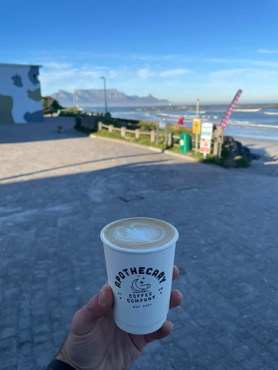 A hand holds an Apothecary Coffee takeaway cup at Big Bay, Table Mountain and the surf blurred behind