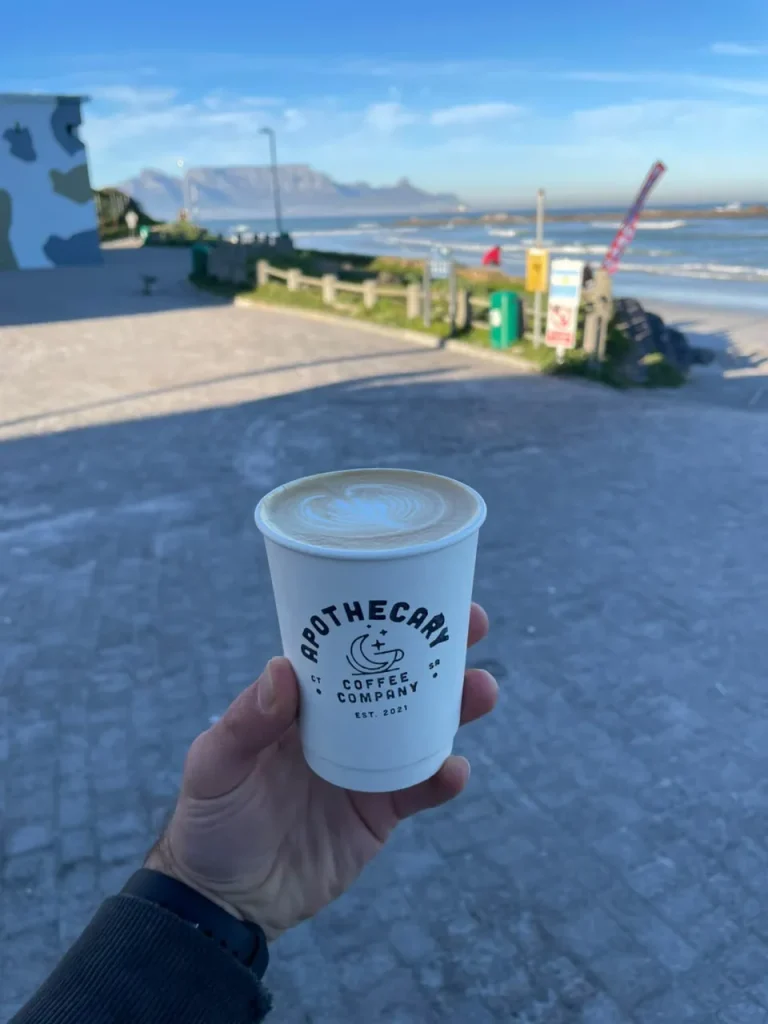 A hand holds an Apothecary Coffee Company takeaway cup topped with swan latte art at the Big Bay lifesaving club, with Table Mountain blurred across the bay