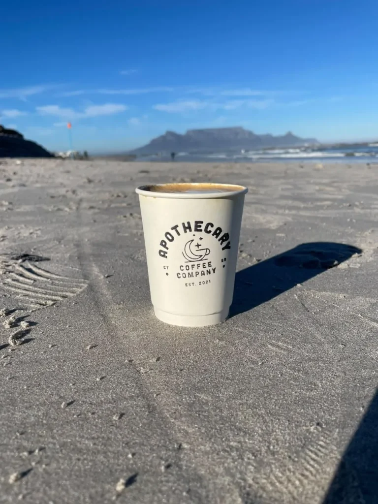 A white Apothecary Coffee Company takeaway cup stands in the beach sand with Table Mountain blurred across Table Bay under a bright blue sky